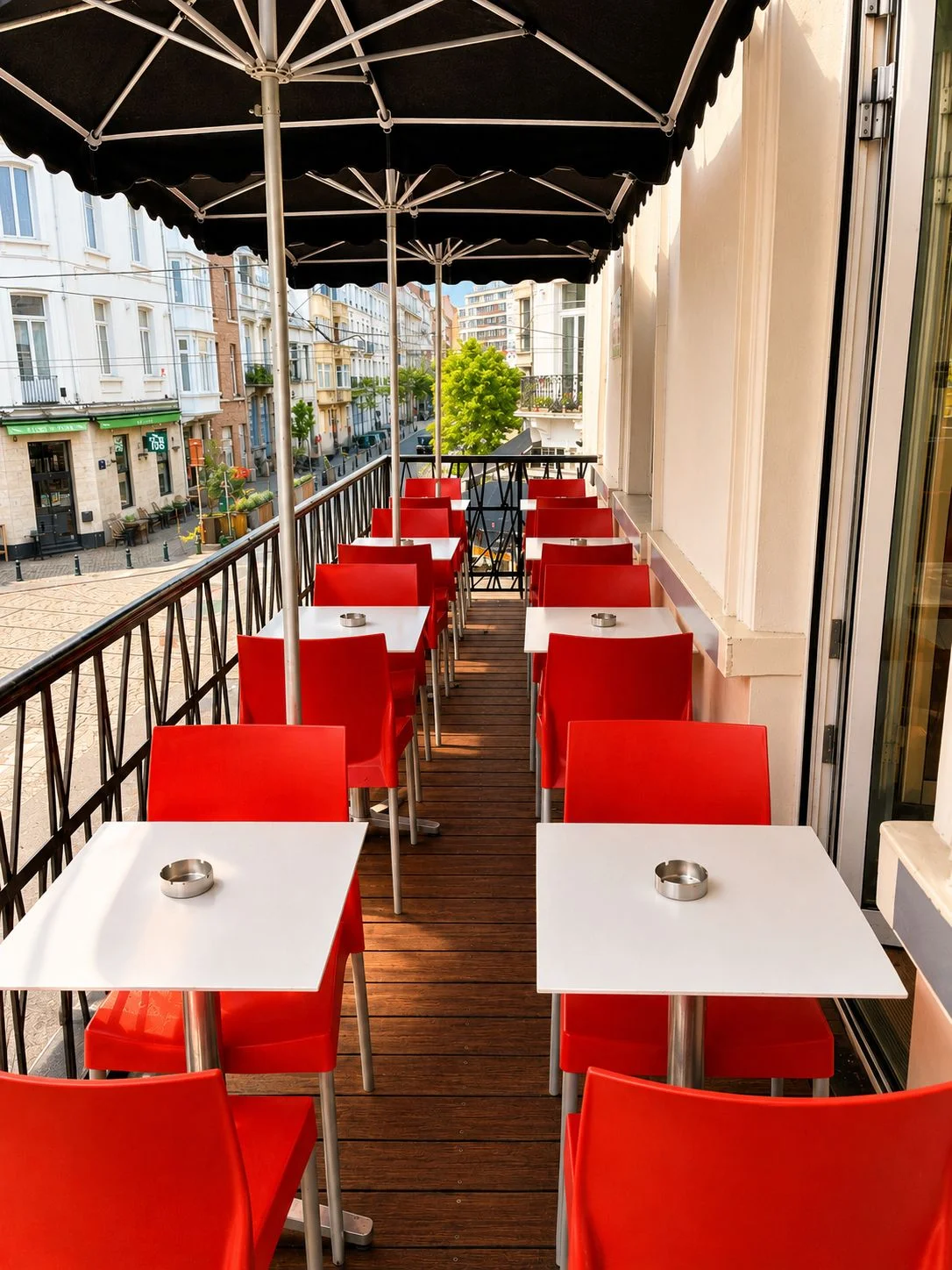 Terrasse-balcon à l'étage du ROXI, tables blanches et chaises rouges sous parasol, vue sur le quartier du Châtelain